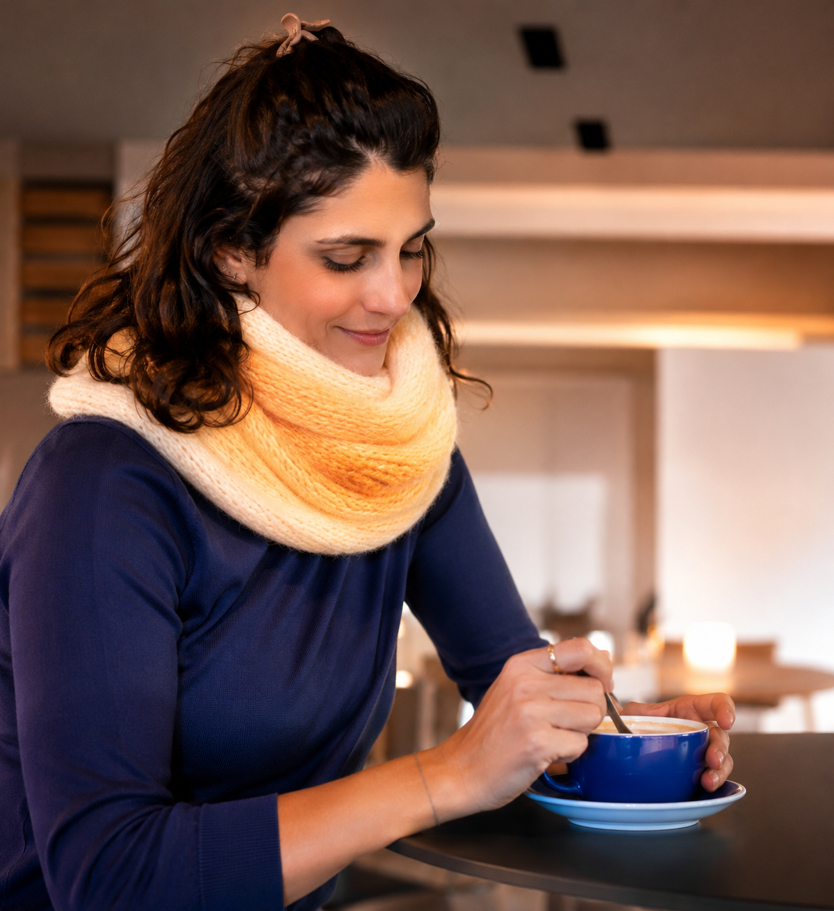 Mulher com cachecol colorido e vestido azul segurando chávena azul numa mesa