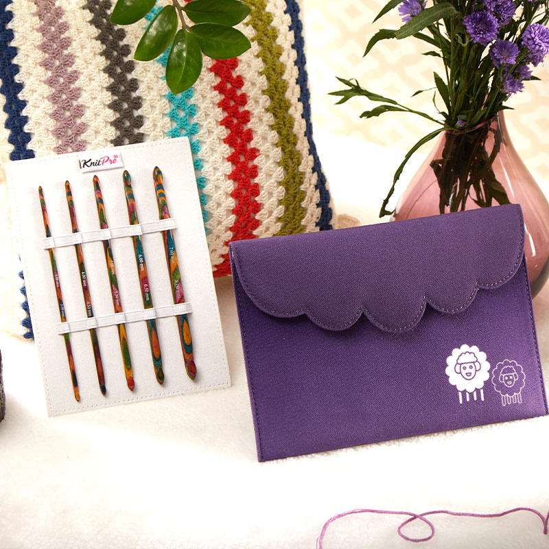 Conjunto de agulhas de crochê em madeira colorida da marca KnitPro junto a carteira roxa com desenhos de ovelhas brancas e lilases, almofada colorida e vaso com flores roxas