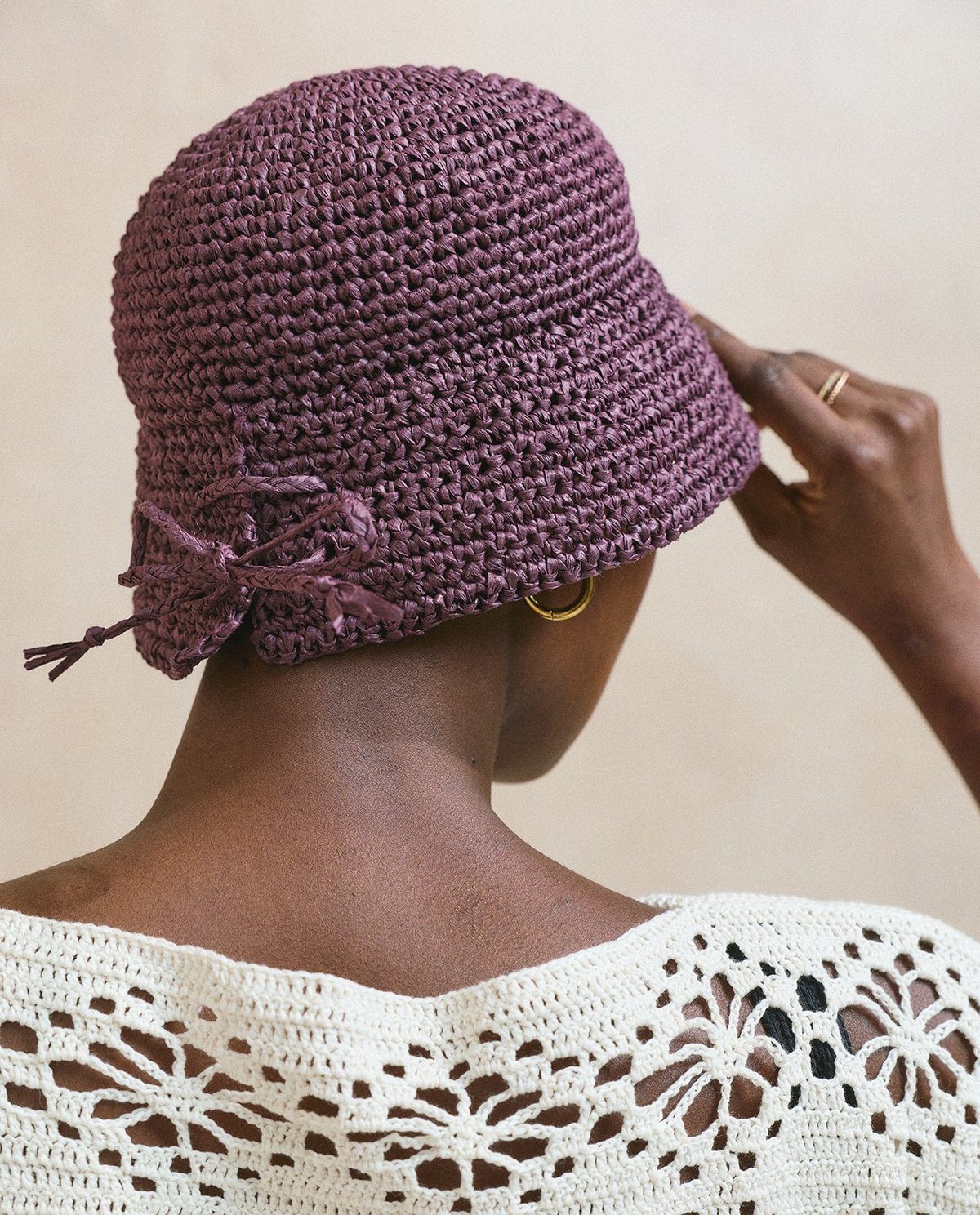 Chapéu de crochet roxo com laço visto de trás, em cabeça de pessoa com pele morena e roupa de crochet branca.