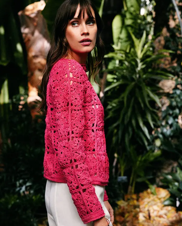 Mulher com camisola de croché cor-de-rosa e calças brancas em ambiente natural com plantas.