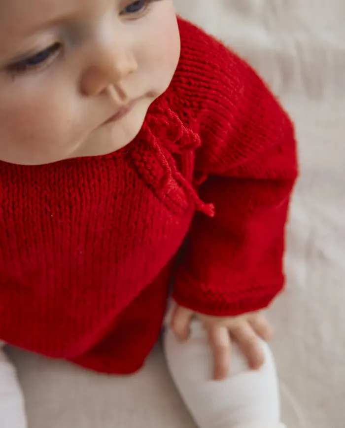 Bebé com camisola vermelha de malha e meia-calça branca num fundo bege claro