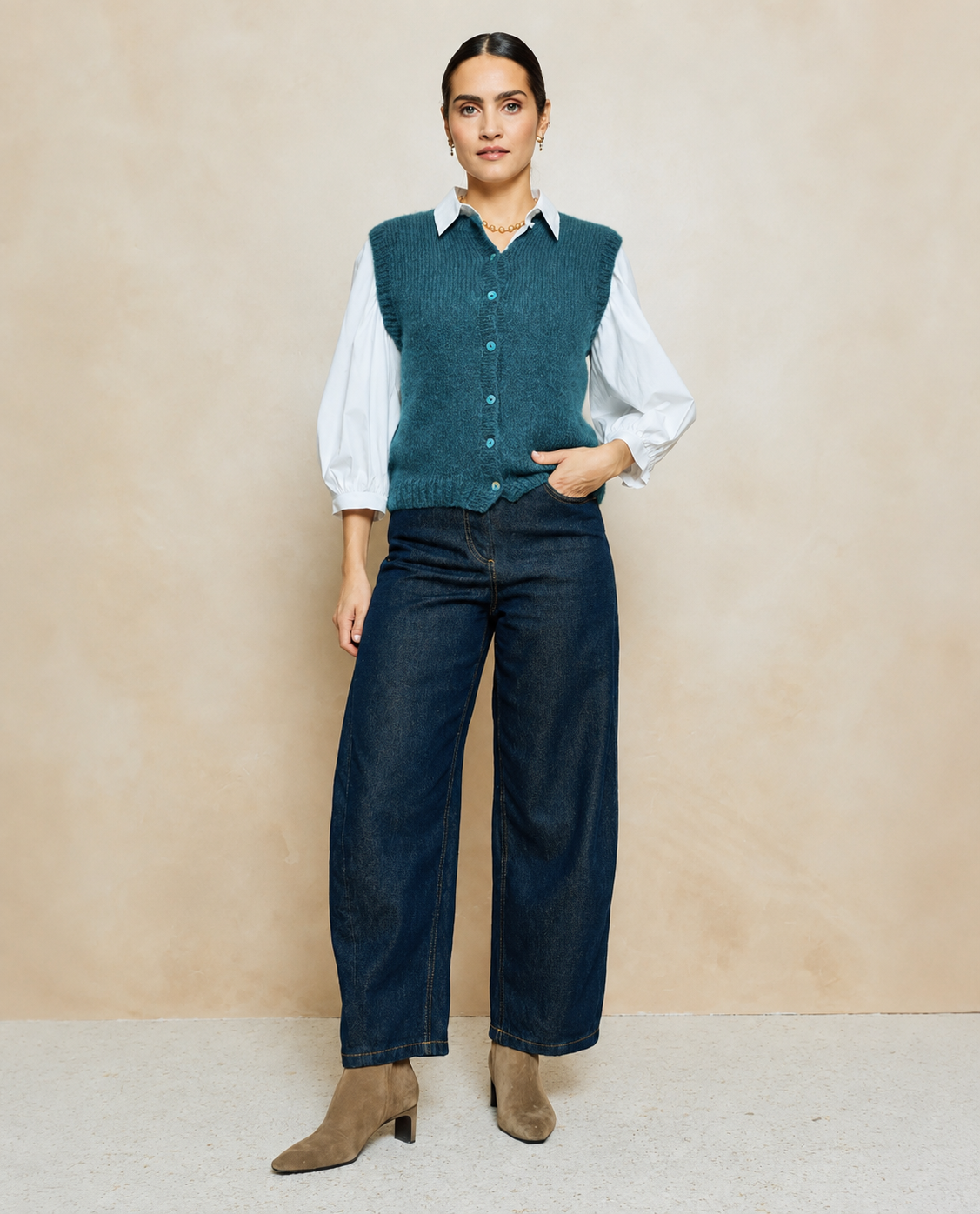 Mulher vestindo camisola azul petróleo sem mangas com botões, camisa branca de manga comprida, calças largas de ganga azul escuro e botins bege.