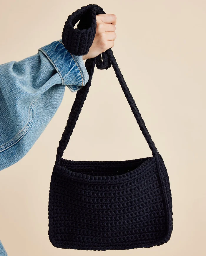 Mala preta de crochet com alça longa segurada por mão vestindo camisa azul de ganga