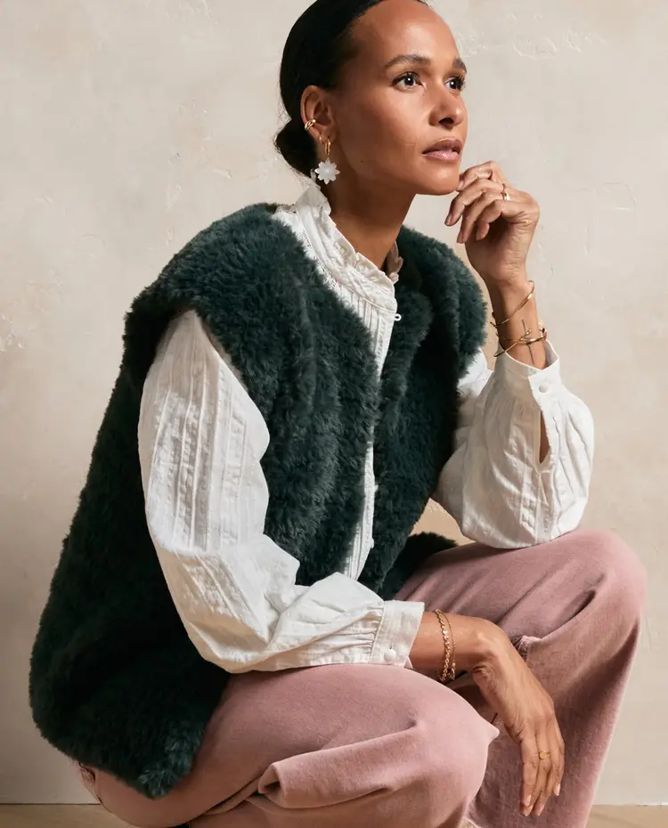 Mulher com colete peludo verde escuro, camisa branca texturada e calças cor-de-rosa, acessórios dourados e brinco branco