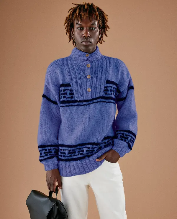 Homem veste pulôver azul com padrão geométrico e botões dourados, calças brancas, segura mala preta