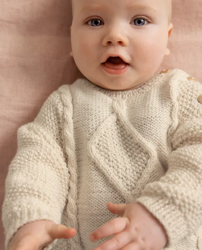 Bebé deitado veste roupa creme de malha grossa com padrão trançado e botões de madeira