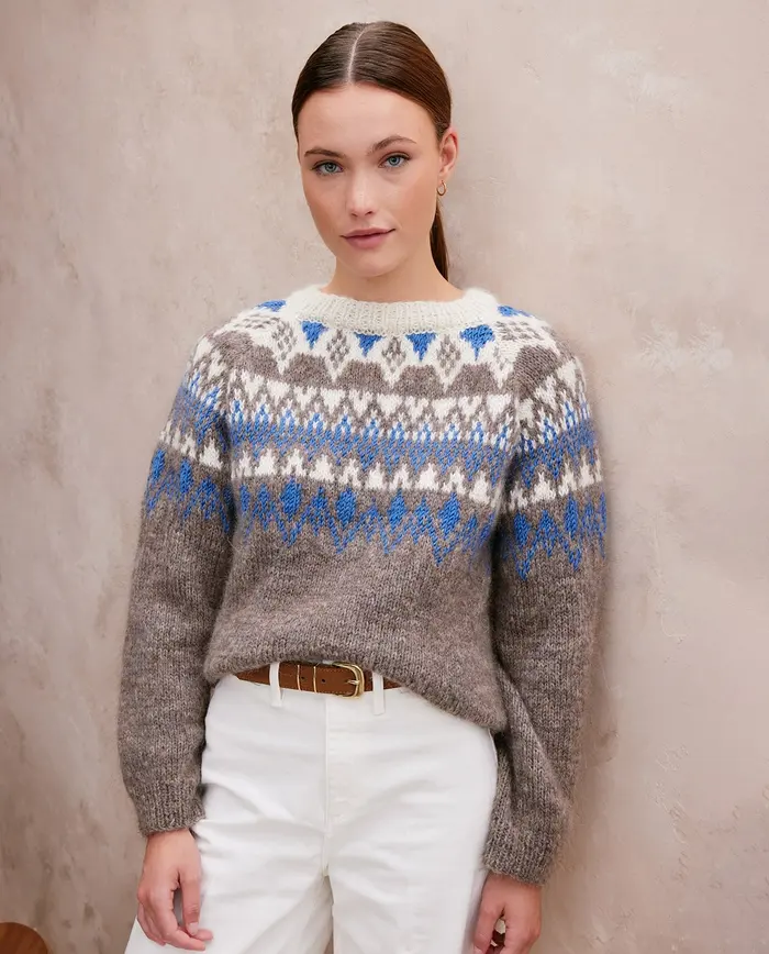 Mulher com camisola de malha cinza com padrão branco e azul e calças brancas.