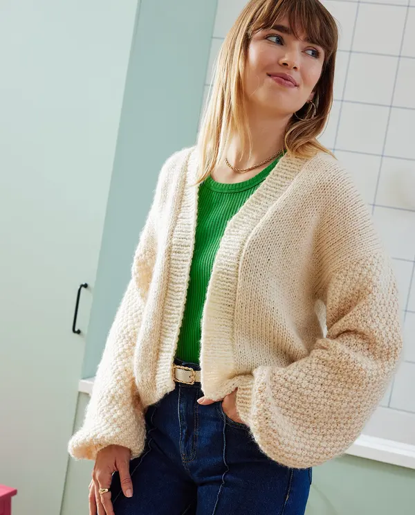 Mulher com camisola de malha bege e camisola verde, jeans, em cozinha com azulejos brancos