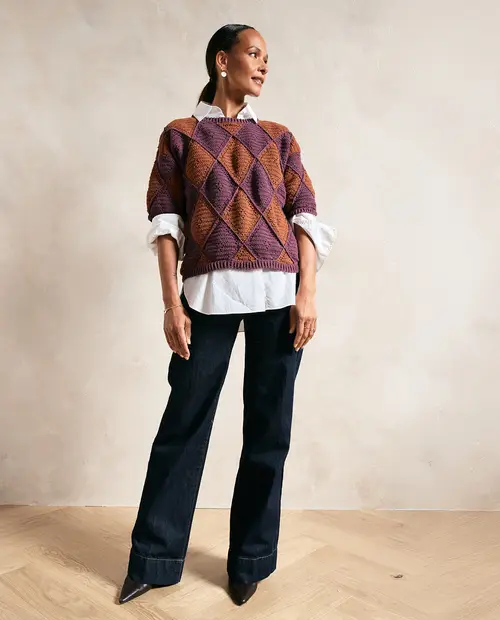 Mulher com camisola de malha em padrões de losangos castanhos e roxos e calças pretas.