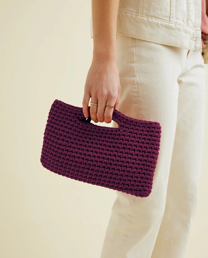 Mala de mão roxa tricotada sendo segurada por pessoa com calças brancas