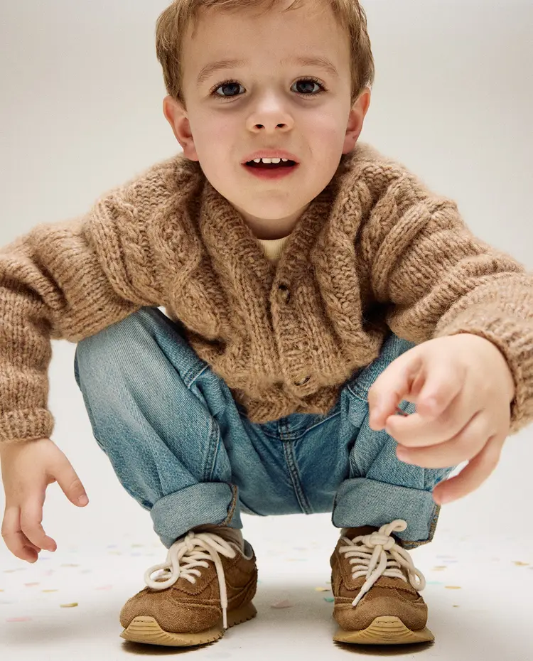 Menino de suéter castanho, jeans e ténis castanhos agachado num chão claro