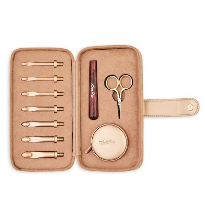 Estojo castanho claro com sete agulhas de croché douradas, tesoura pequena dourada, marcador de madeira e estojo redondo.