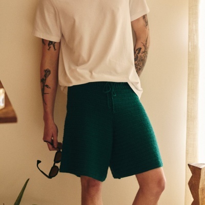 Homem com calções verdes de malha e t-shirt branca, segurando óculos de sol.