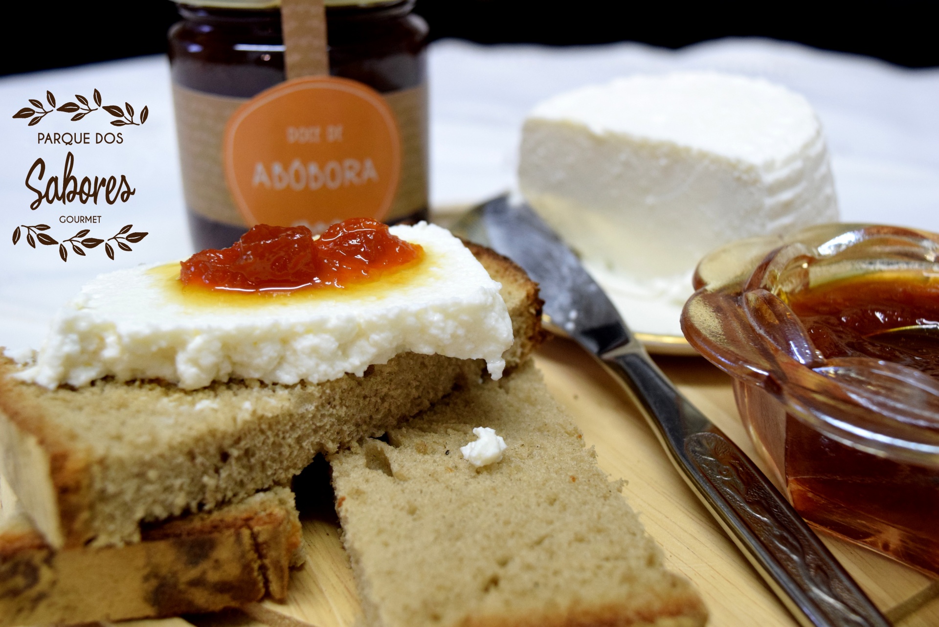 Pão com queijo cremoso e doce de abóbora em tábua de madeira com frasco e bloco de queijo ao fundo