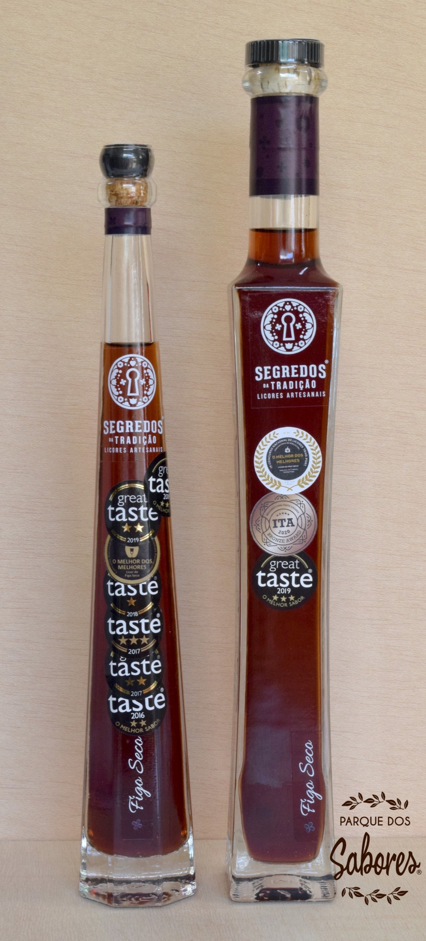 Duas garrafas de licor artesanal Segredos da Tradição com etiquetas de prémios, em vidro transparente com tampas diferentes, sobre fundo de madeira clara