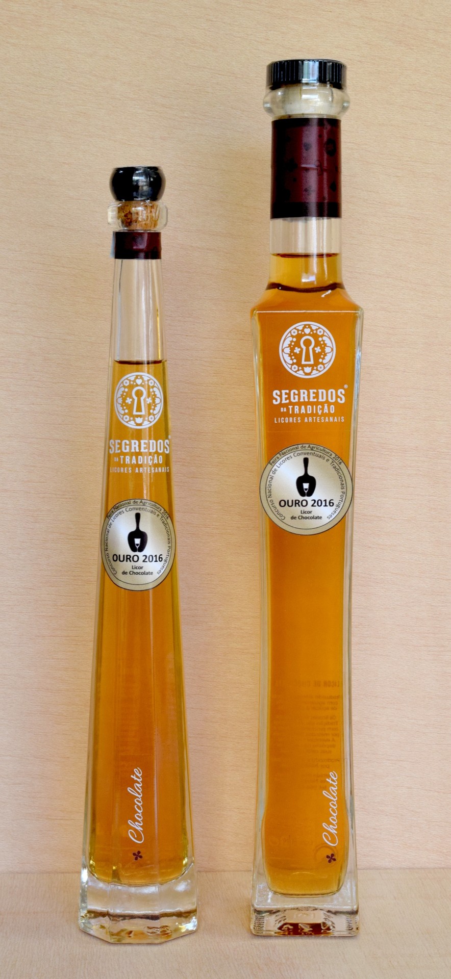 Duas garrafas altas de licor artesanal âmbar com rótulos SEGREDOS DA TRADIÇÃO e OURO 2016