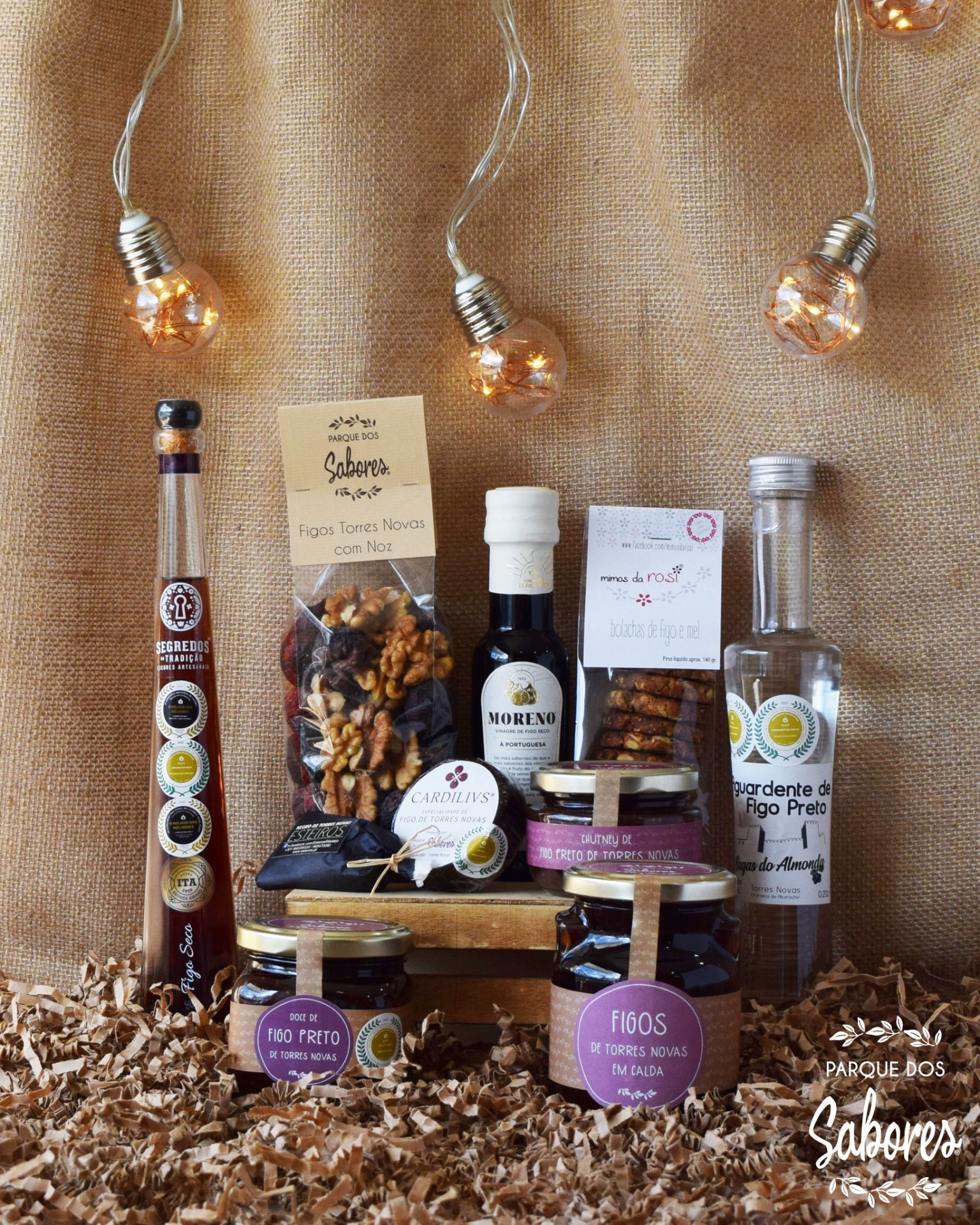 Produtos alimentares variados em frascos e embalagens com figos secos, azeite e doces, fundo tecido castanho e luzes decorativas