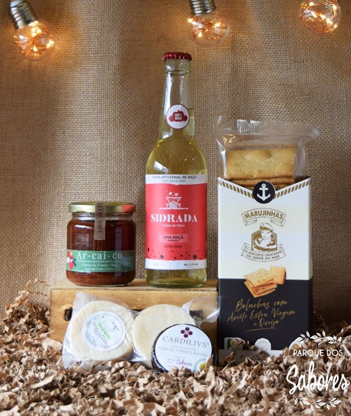 Produtos gourmet incluindo compota, sidra, bolachas e queijos sobre fundo castanho com luzes decorativas