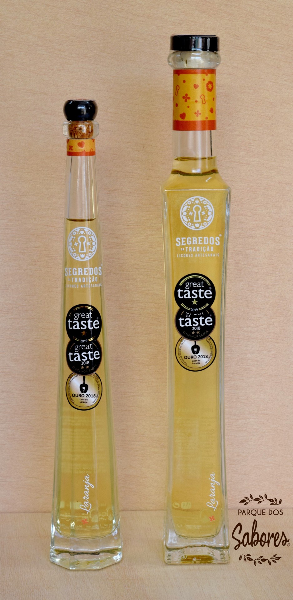 Duas garrafas transparentes de licor amarelo claro da marca Segredos de Tradição com rótulos e tampas pretas