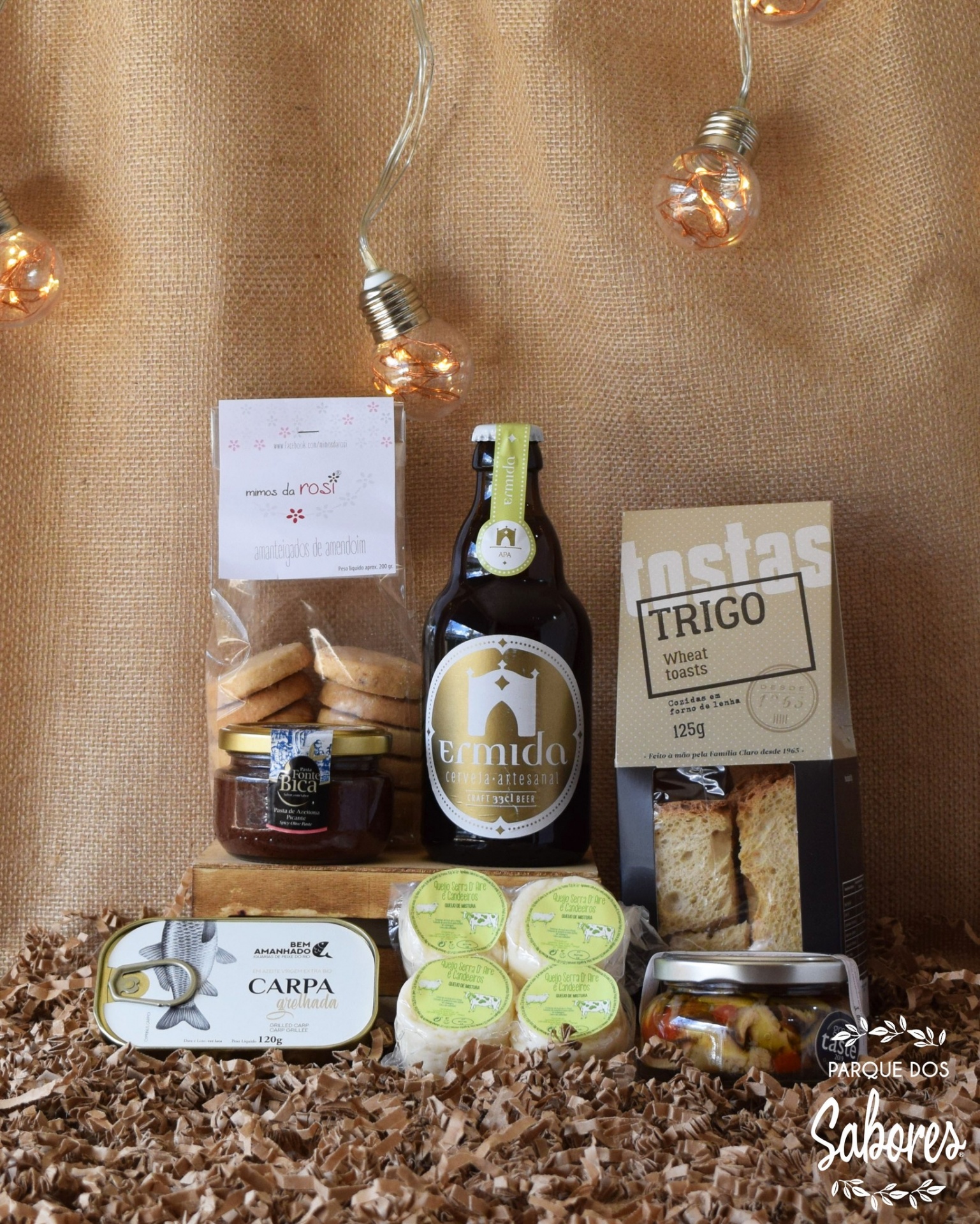 Produtos gourmet variados incluindo cerveja, queijos, tostas, biscoitos e conservas sobre papel amassado com luzes decorativas