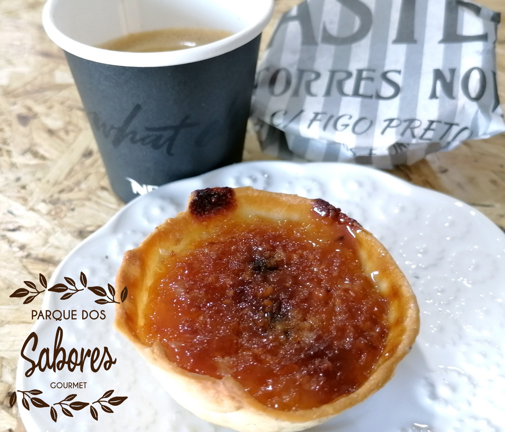 Pastel de Feijão de Torres Novas Tarteleta caramelizada num prato branco, chávena preta com bebida e embalagem de papel com texto