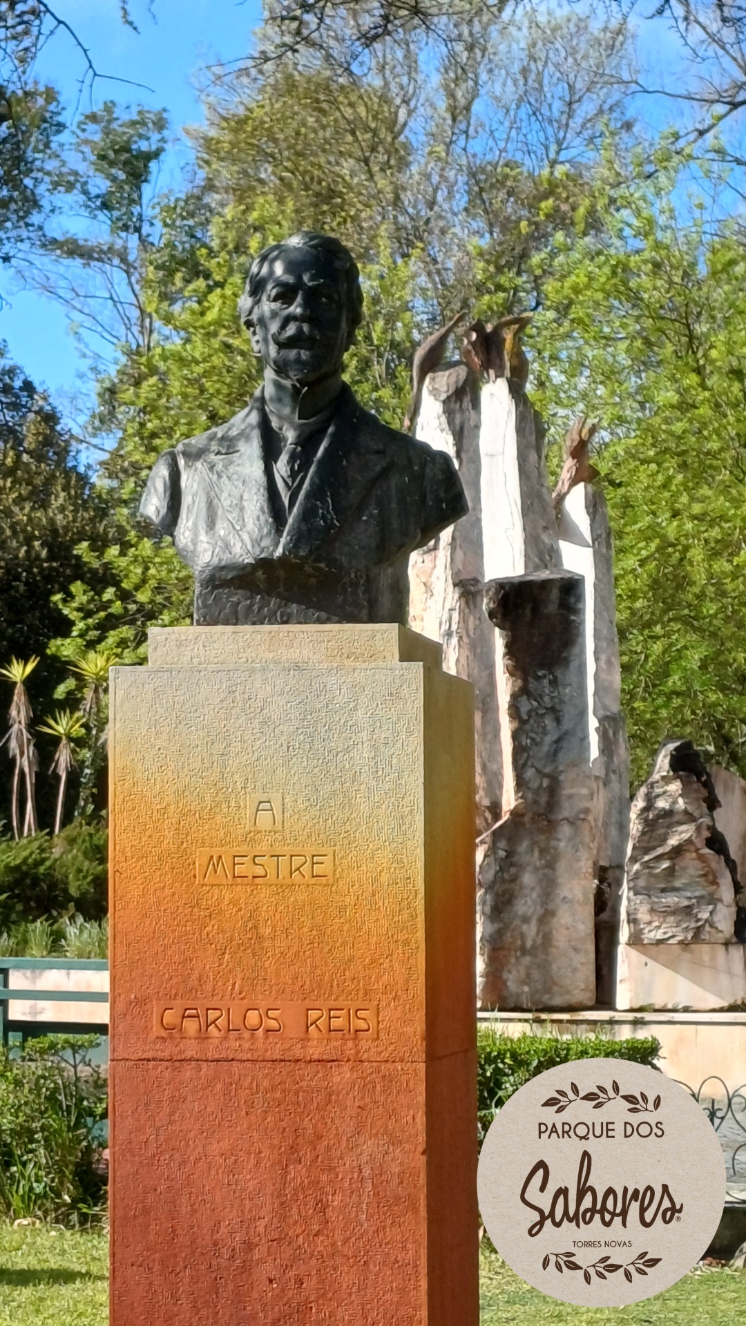 Busto em bronze de Carlos Reis sobre pedestal com texto Ai Mestre Carlos Reis num parque com esculturas e logo Parque dos Sabores