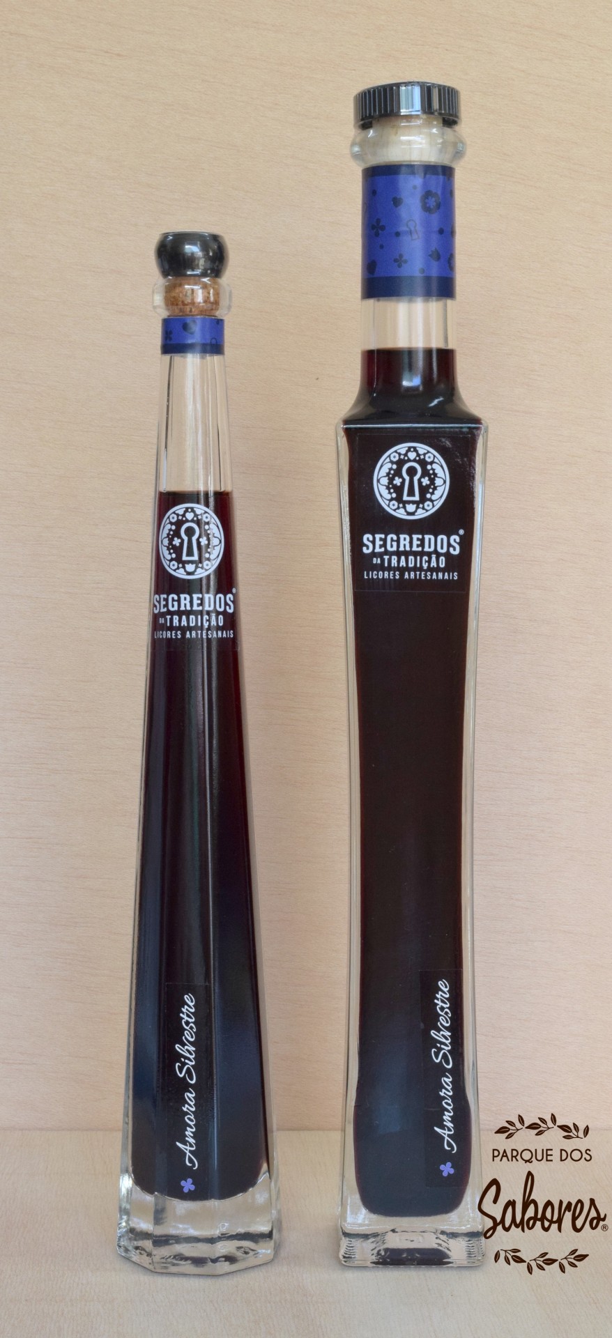 Duas garrafas de licor artesanal em vidro com etiquetas azuis e texto branco, sobre fundo de madeira clara