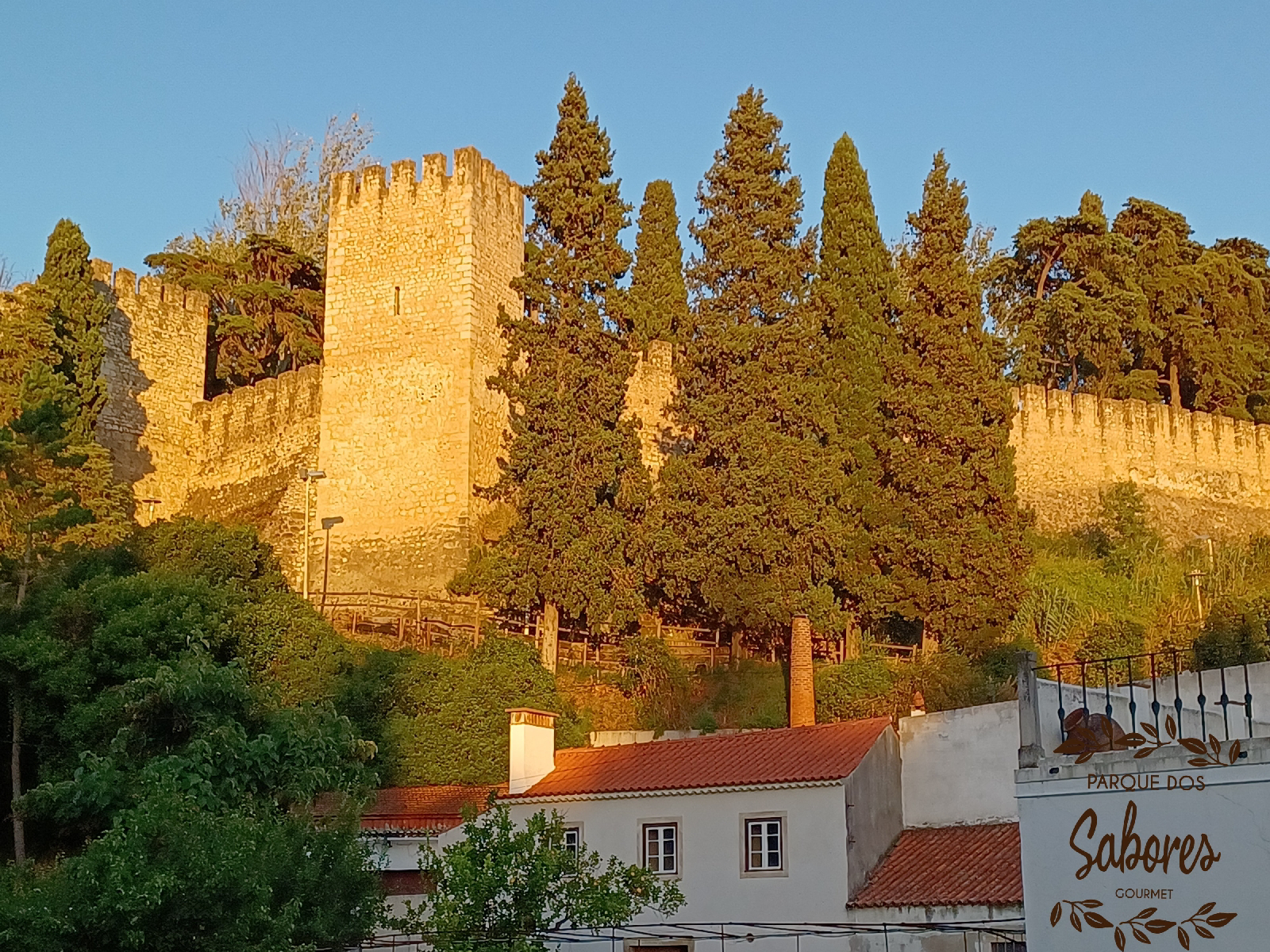 Muralha medieval em pedra com torres, árvores e casas brancas com telhados vermelhos.