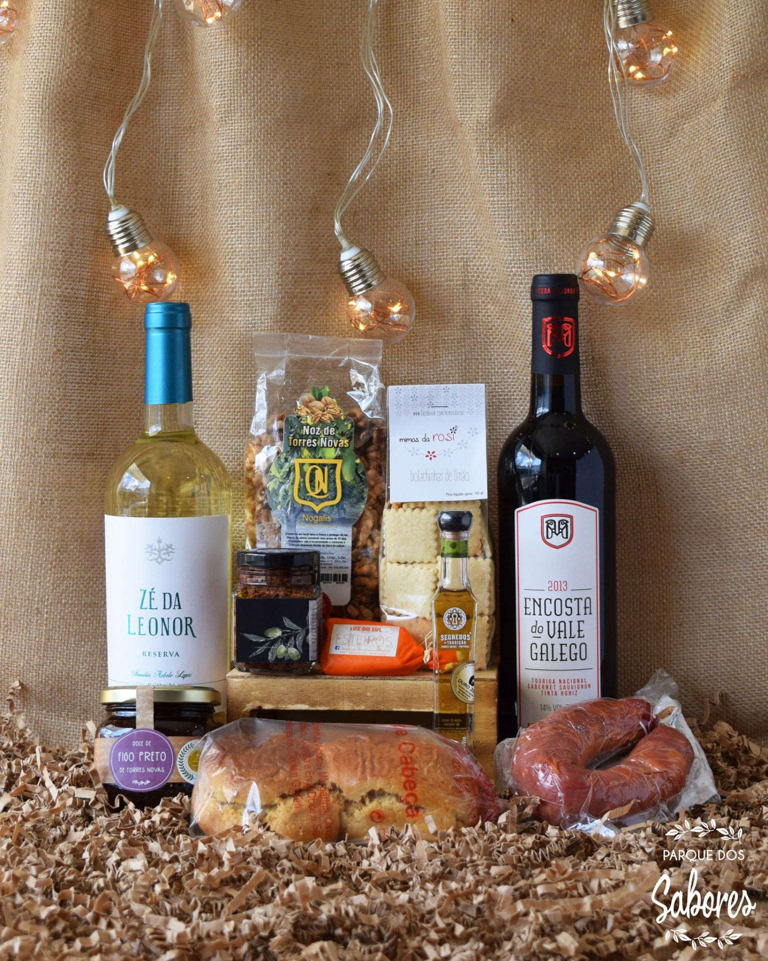 Conjunto de produtos gourmet com vinhos, nozes, queijo, patê, azeite, broa e chouriço sobre fundo de juta com luzes decorativas