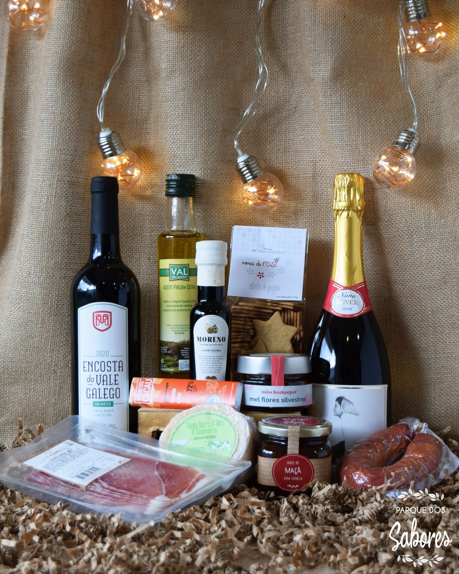 Produtos gourmet incluindo vinho, espumante, azeite, embutidos e queijo em fundo tecido castanho com luzes decorativas