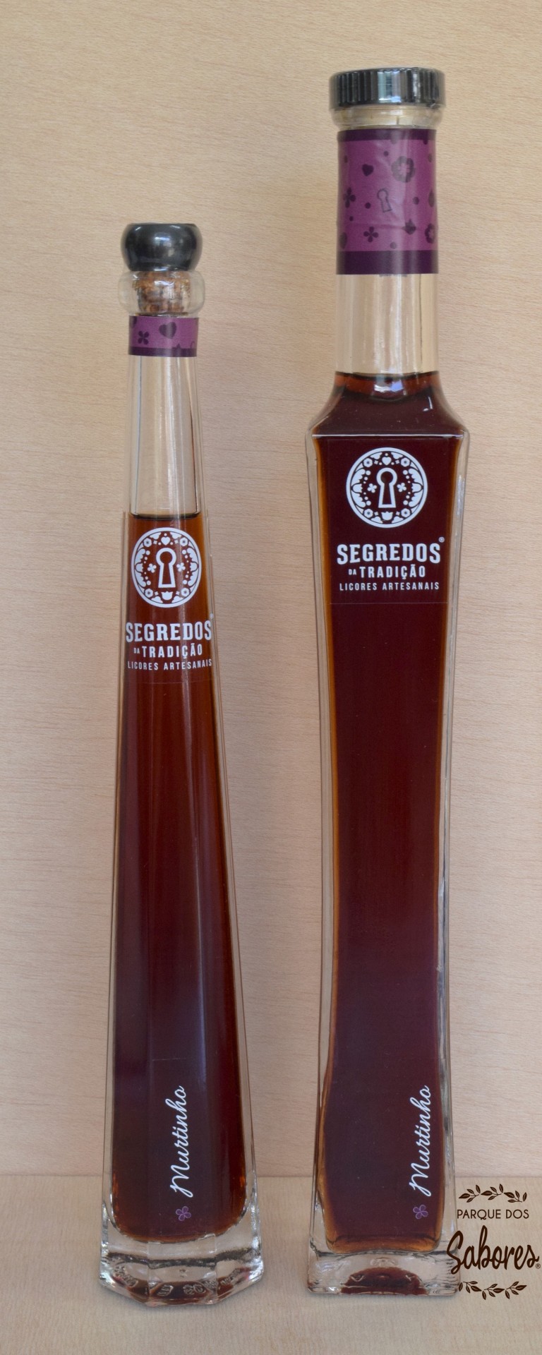 Duas garrafas de licor castanho com etiquetas brancas e tampas diferentes sobre fundo madeira clara