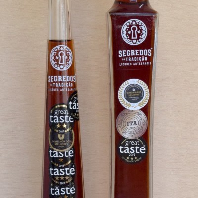 Duas garrafas de licor artesanal Segredos da Tradição com etiquetas de prémios, em vidro transparente com tampas diferentes, sobre fundo de madeira clara