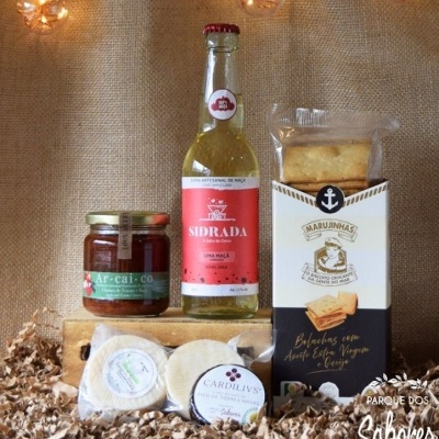 Produtos gourmet incluindo compota, sidra, bolachas e queijos sobre fundo castanho com luzes decorativas