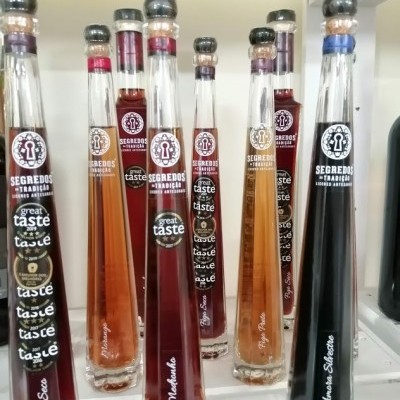 Garrafas de vidro estreitas com líquidos coloridos e rótulos da marca Segredos da Tradição em prateleira