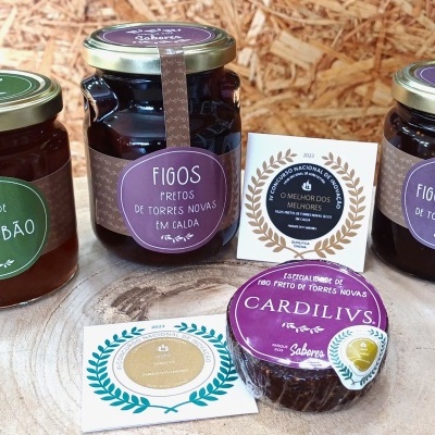 Três frascos de compota com tampas e etiquetas coloridas em fundo de cortiça, acompanhados por pequenos cartões decorativos.