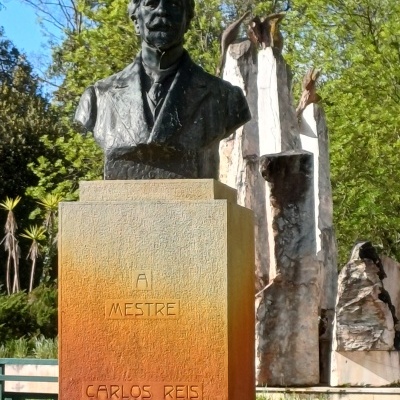 Busto em bronze de Carlos Reis sobre pedestal com texto Ai Mestre Carlos Reis num parque com esculturas e logo Parque dos Sabores