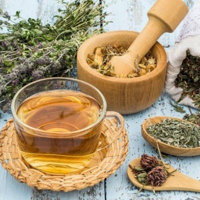 Chávena de chá com ervas secas, almofariz de madeira e saco branco com renda sobre mesa azul de madeira.