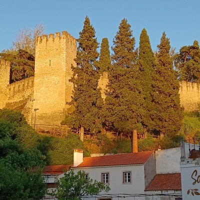 Muralha medieval em pedra com torres, árvores e casas brancas com telhados vermelhos.