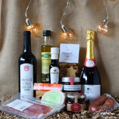 Produtos gourmet incluindo vinho, espumante, azeite, embutidos e queijo em fundo tecido castanho com luzes decorativas