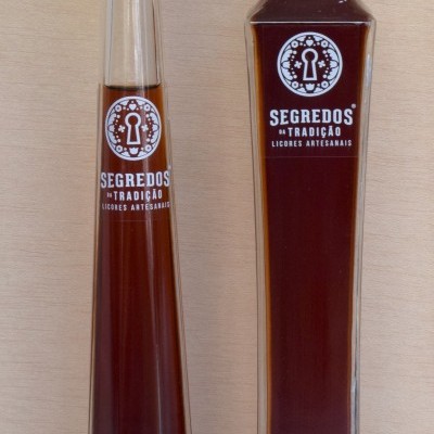 Duas garrafas de licor castanho com etiquetas brancas e tampas diferentes sobre fundo madeira clara