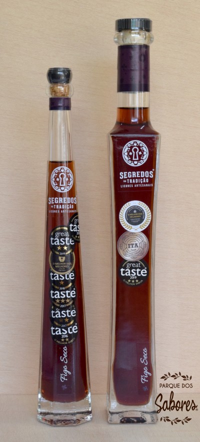 Duas garrafas de licor artesanal Segredos da Tradição com etiquetas de prémios, em vidro transparente com tampas diferentes, sobre fundo de madeira clara