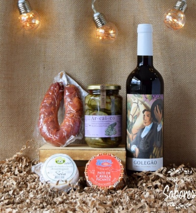 Produtos gourmet incluindo chouriço, picles, queijo, paté e vinho tinto dispostos em caixa de madeira sobre lascas de madeira e fundo bege