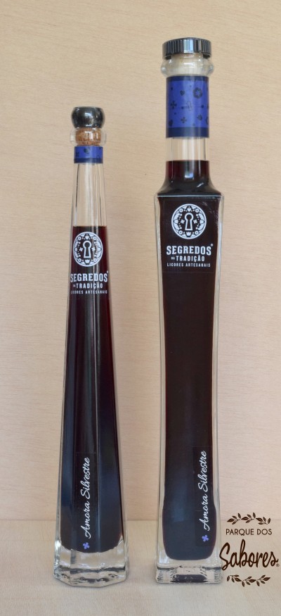 Duas garrafas de licor artesanal em vidro com etiquetas azuis e texto branco, sobre fundo de madeira clara