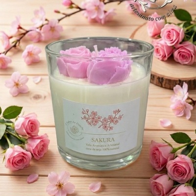 Vela aromática artesanal Sakura em copo de vidro com flores rosas no topo e ramos floridos sobre mesa de madeira.