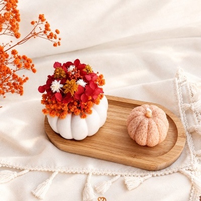 Arranjo decorativo com abóbora branca e flores e vela em forma de abóbora sobre base de madeira.