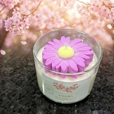 Vela aromática em vidro transparente com topo em forma de flor lilás e etiqueta com flores e texto, sobre superfície escura e fundo de flores cor-de-rosa.