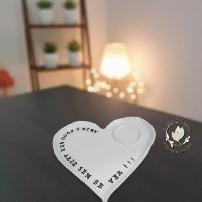 Prato cerâmico branco em forma de coração com texto preto e relevo, em superfície preta, com estante e luz difusa ao fundo.