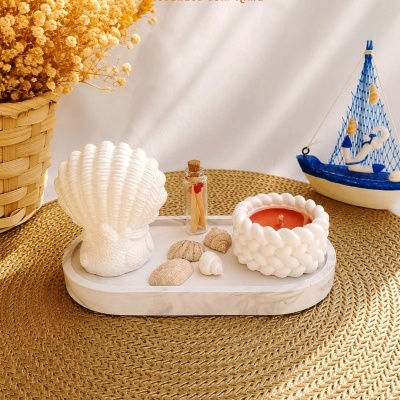 Conjunto decorativo com vela em cera, castiçal concha e conchas naturais sobre tabuleiro branco em superfície trançada.