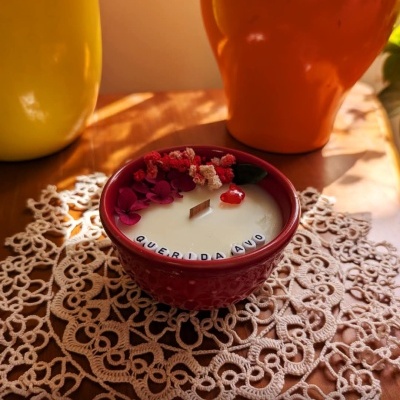Vela branca decorativa numa taça vermelha com flores secas e texto QUERIDA AVÓ sobre renda e vasos coloridos ao fundo.
