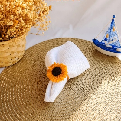 Porta-guardanapo com flor amarela em tecido branco sobre base dourada, arranjo de flores secas em cesto e enfeite de barco azul e branco